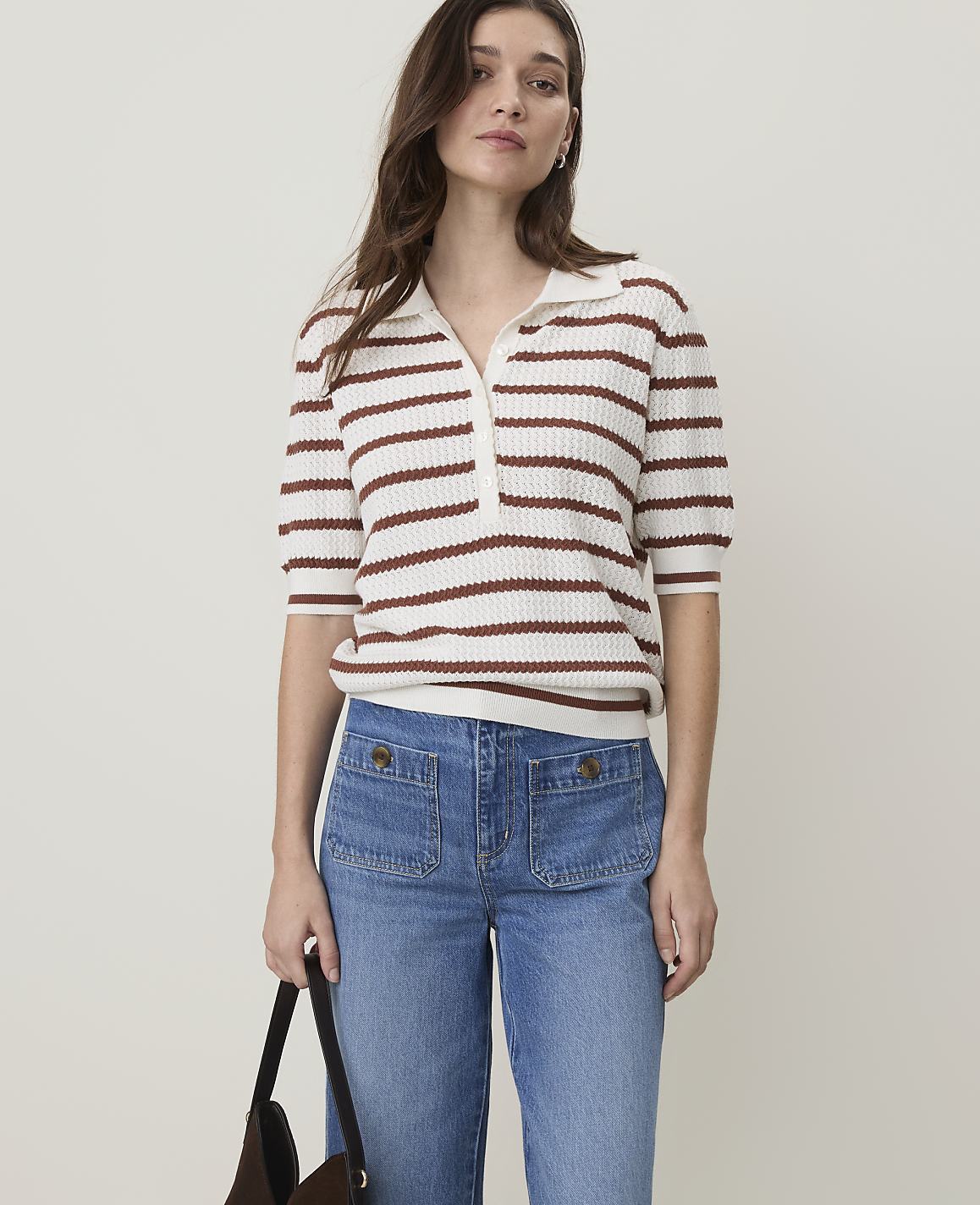 A woman in a striped polo shirt and denim pants stands against a plain white background, holding a dark bag.
