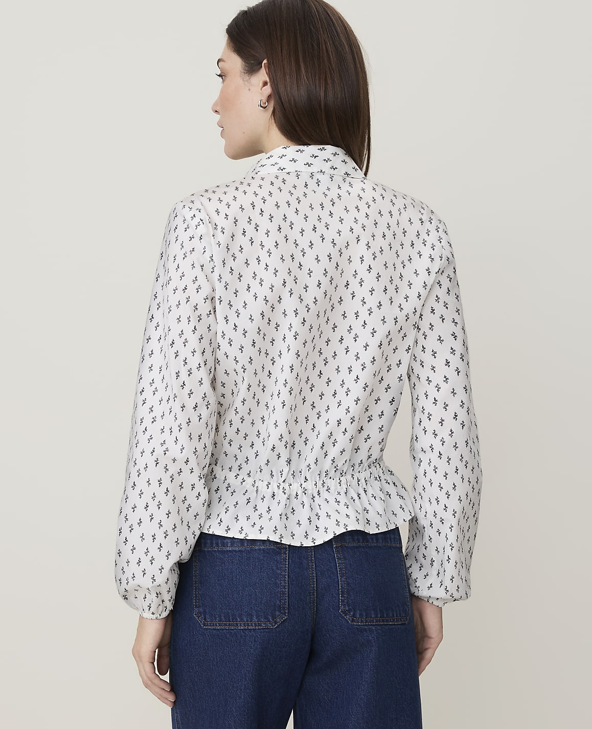 A woman wearing a white patterned blouse and blue jeans stands against a plain, light-colored background.
