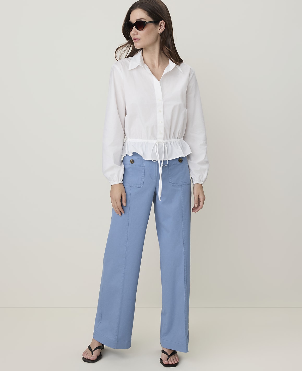 A woman in a white blouse and blue pants stands against a plain background.