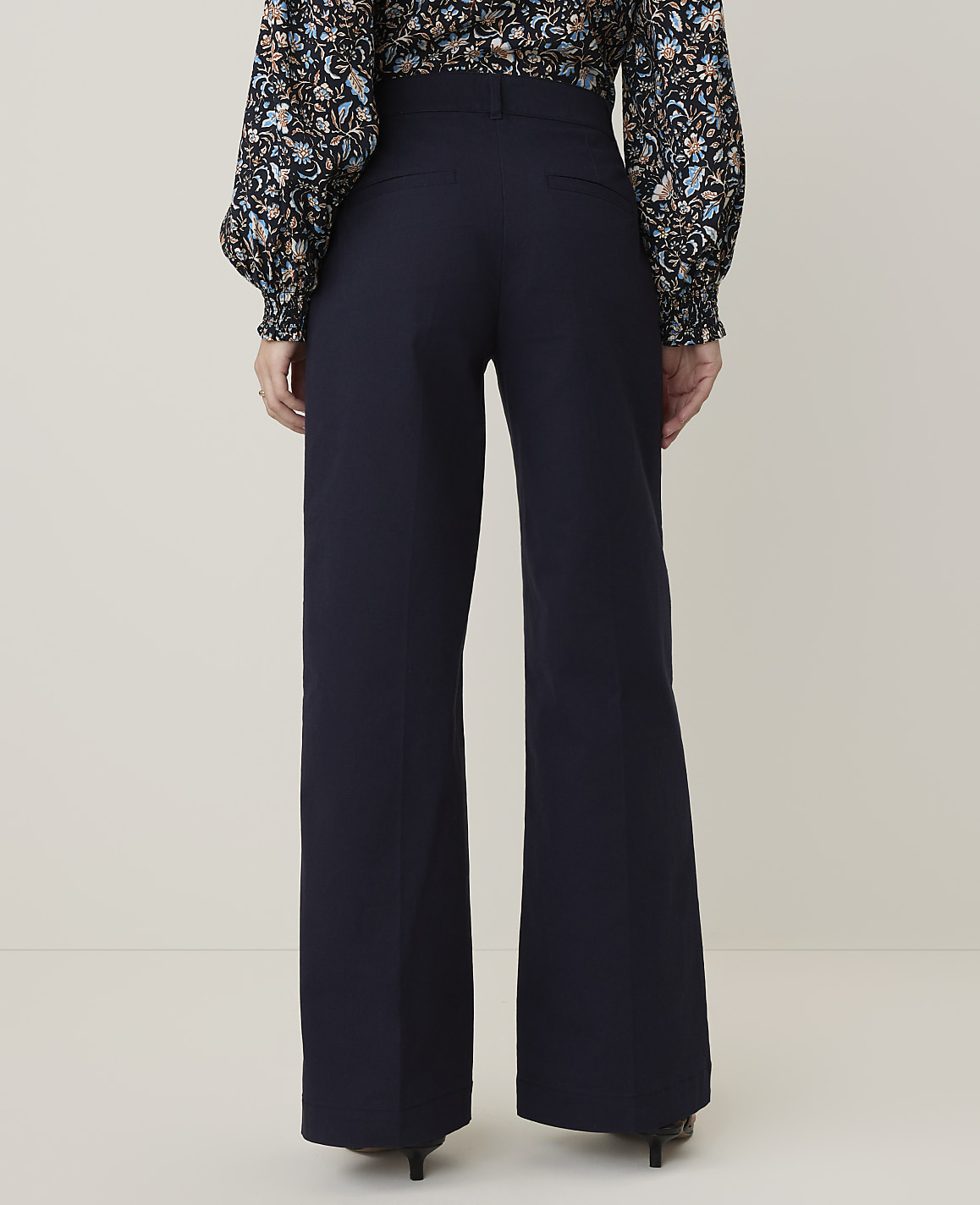A person wearing a floral blouse and dark wide-leg pants stands against a plain, light-colored background.