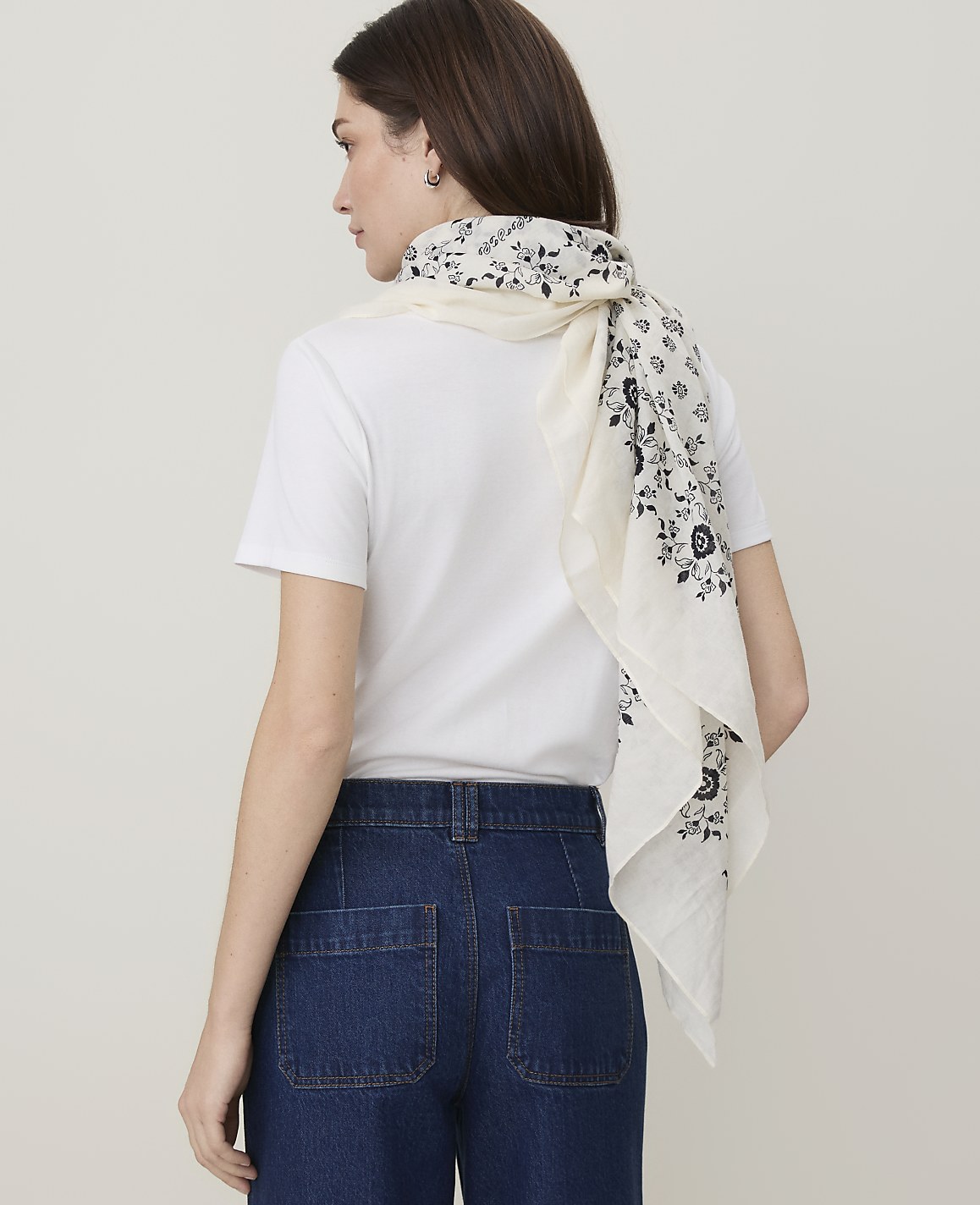 A woman in a white t-shirt and dark denim jeans stands against a plain white background, wearing a floral patterned scarf.
