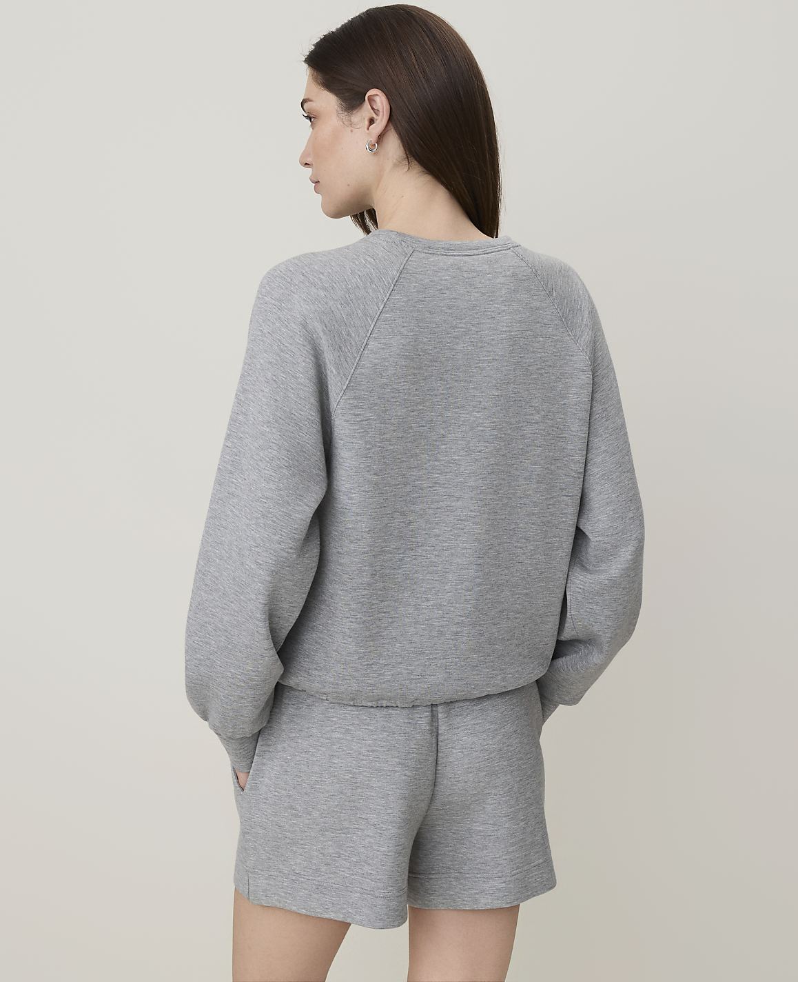 A woman in a grey sweatshirt and shorts stands with her back to the camera against a plain grey background.