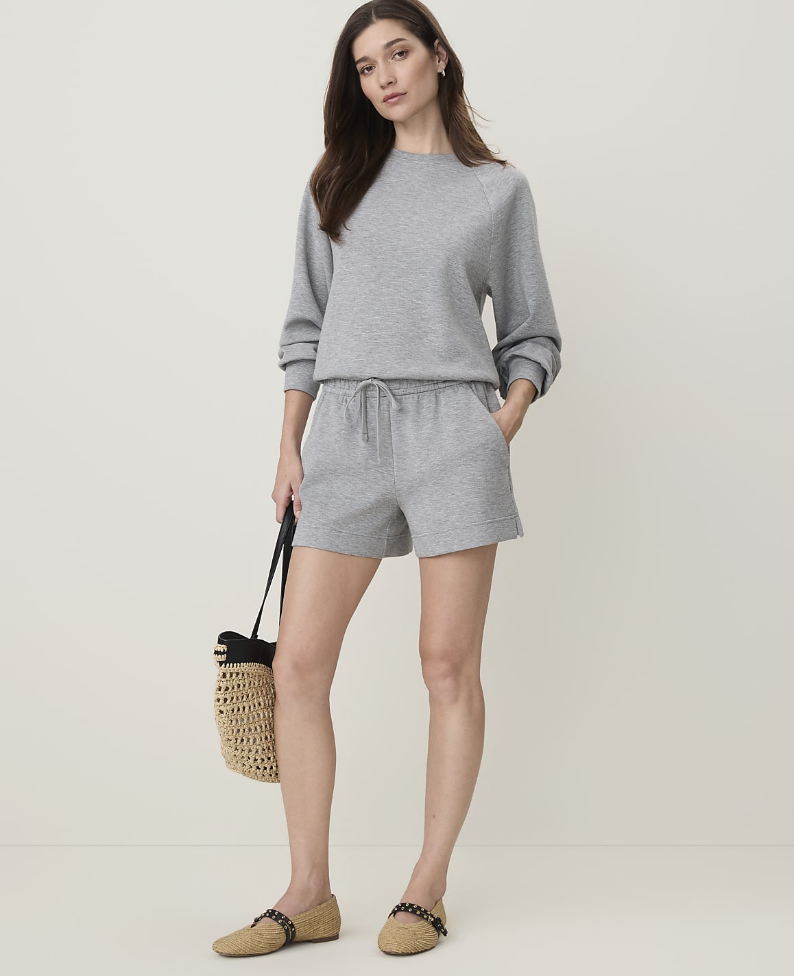A woman in a grey sweatshirt and shorts stands with a straw bag and flats against a plain grey background.