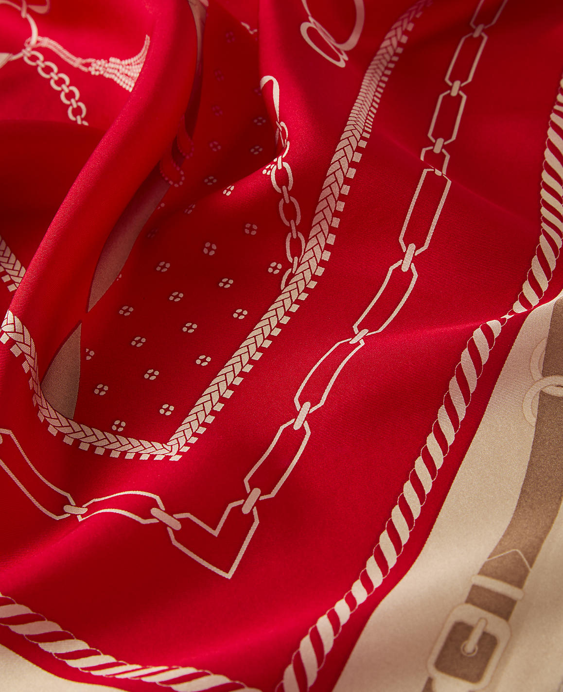 A vibrant red silk scarf with a chain and buckle pattern in the foreground is set against a creamy background with a similar design.