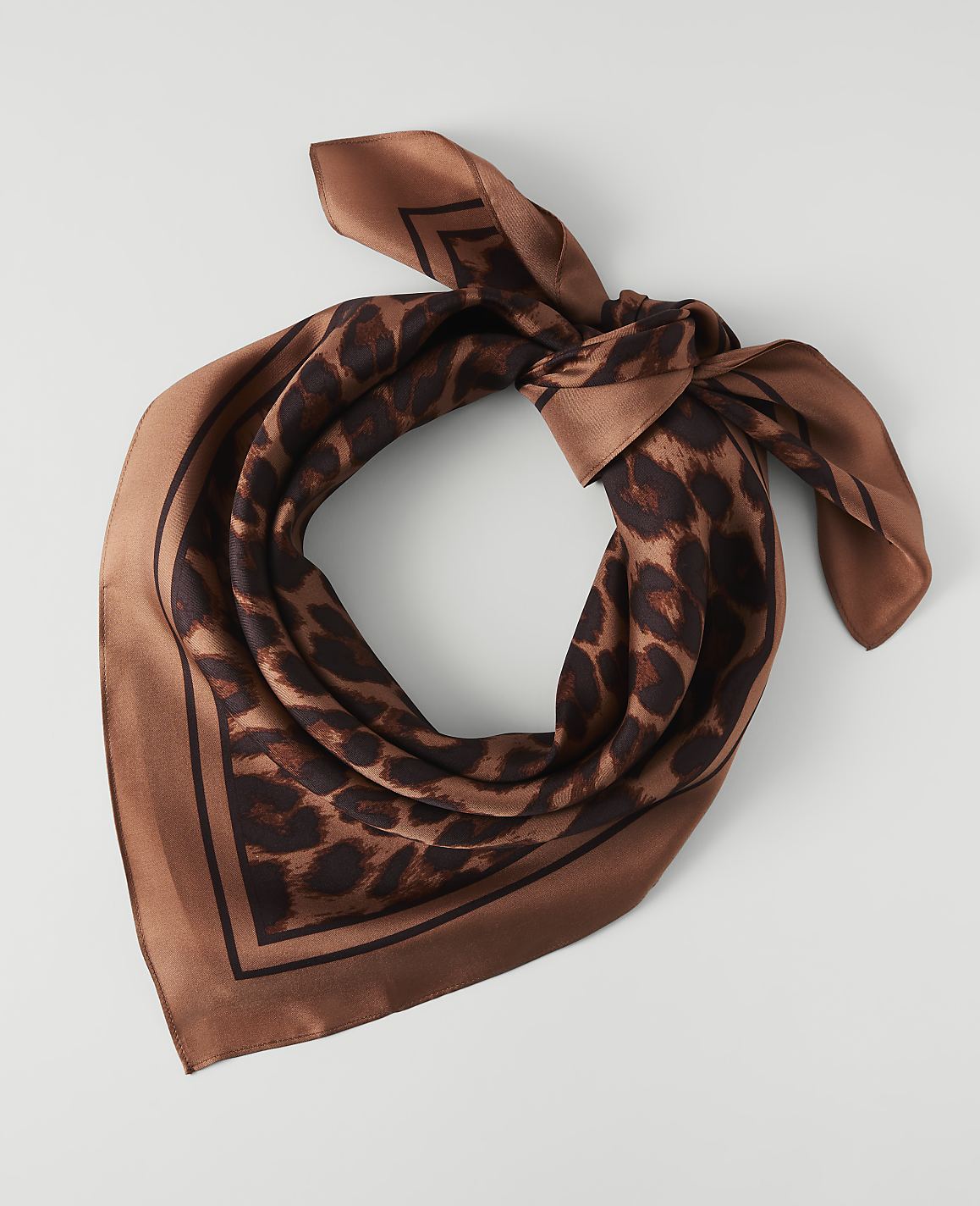 A brown leopard print silk scarf is tied in a knot in the foreground, against a plain white background.