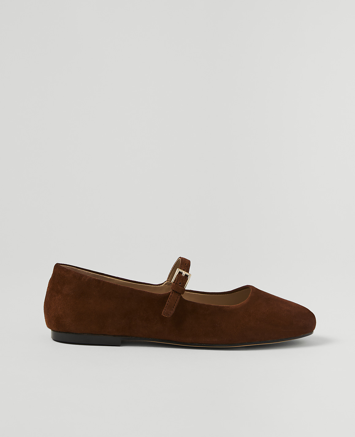 A brown suede Mary Jane flat shoe with a buckle strap rests on a plain white background.