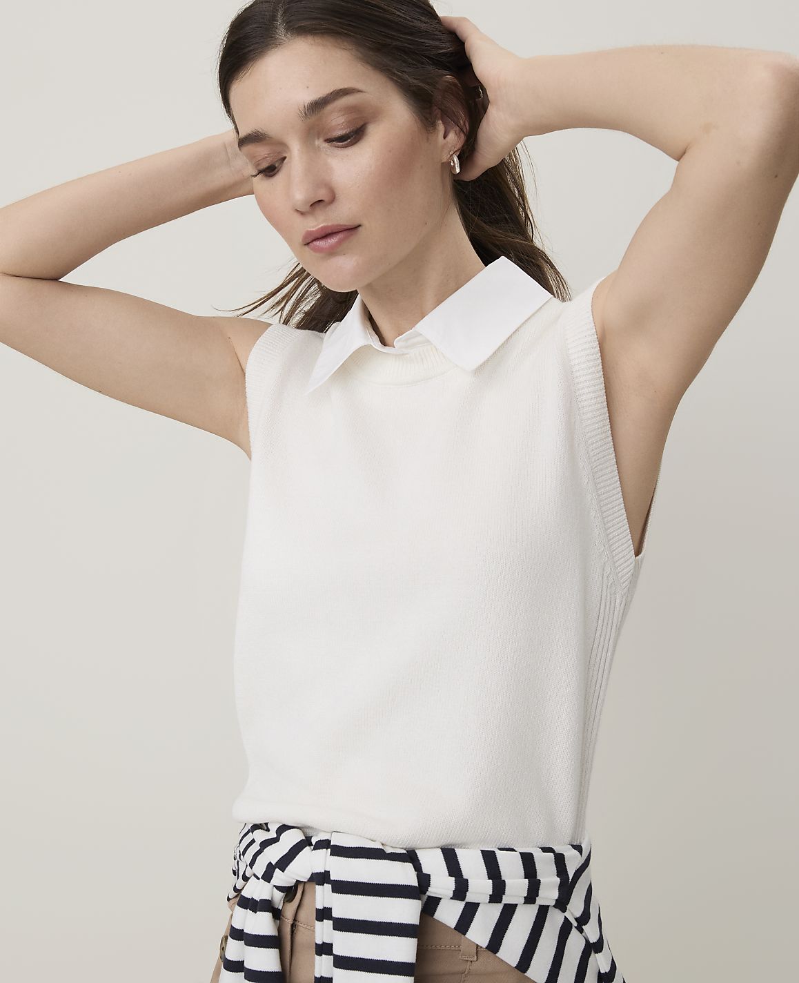 A woman with her hands behind her head wears a white sleeveless collared top and a striped sweater tied around her waist against a plain, light background.