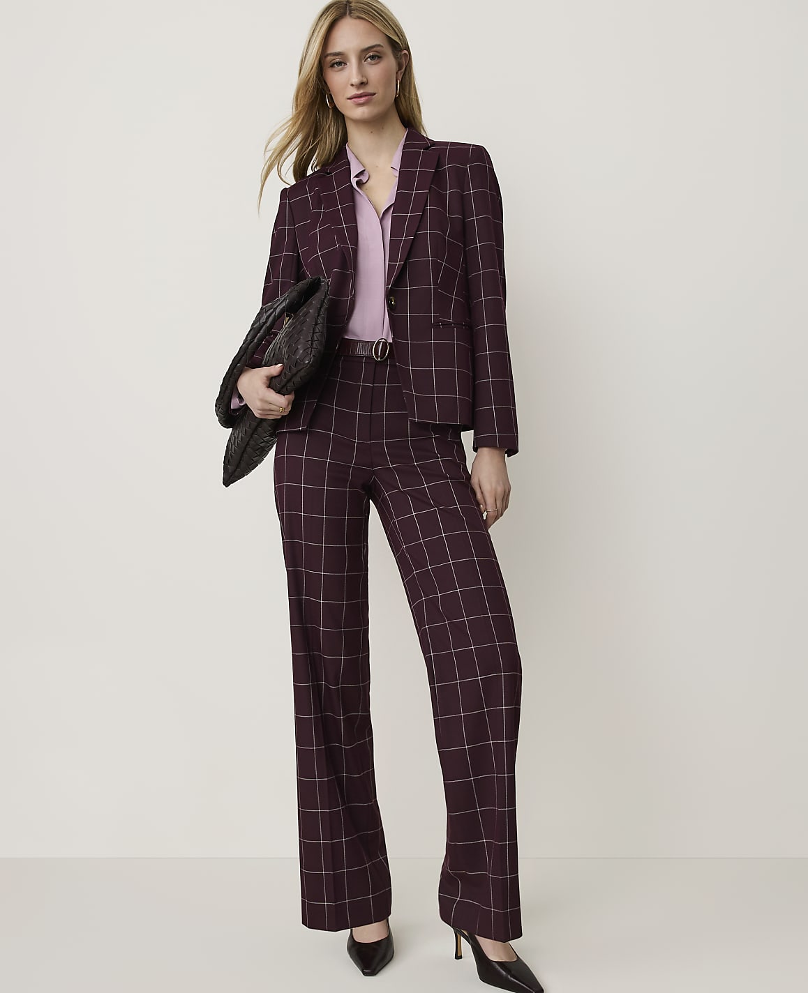 A woman in a burgundy plaid suit and pink blouse stands against a plain, light-colored background, holding a black clutch.