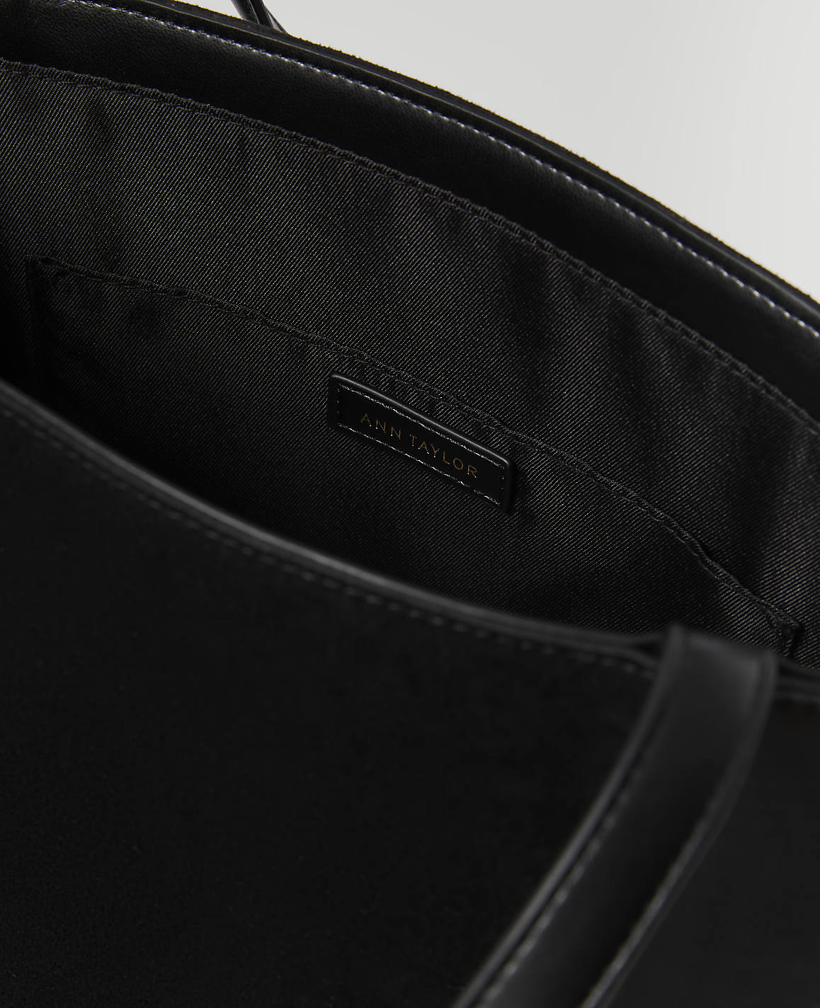 A black Ann Taylor tote bag is shown with its interior lining and a small pocket visible in the foreground, against a dark, out-of-focus background.