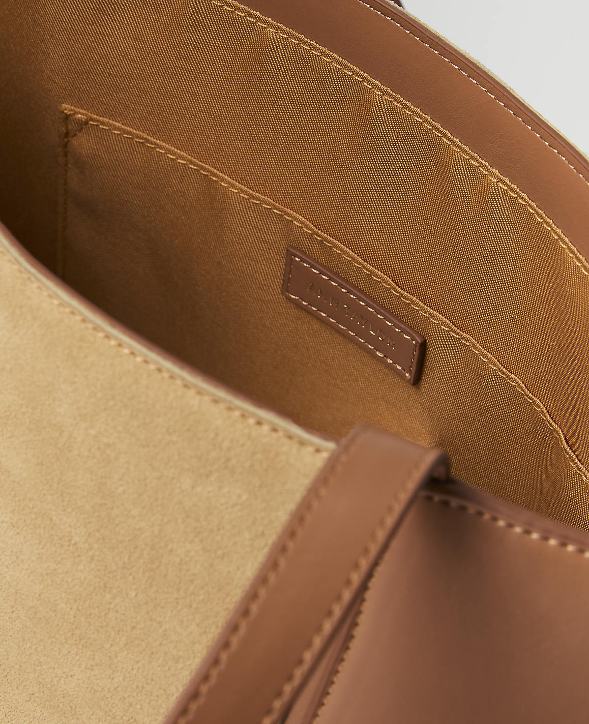 A close-up view of a tan suede tote bag's interior with brown leather trim and straps in the foreground, and a small brand tag visible in the background.