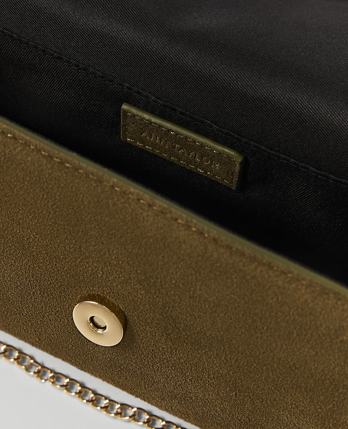 A close-up of an olive green suede clutch with a gold chain strap in the foreground, and a black interior with a brand tag in the background.