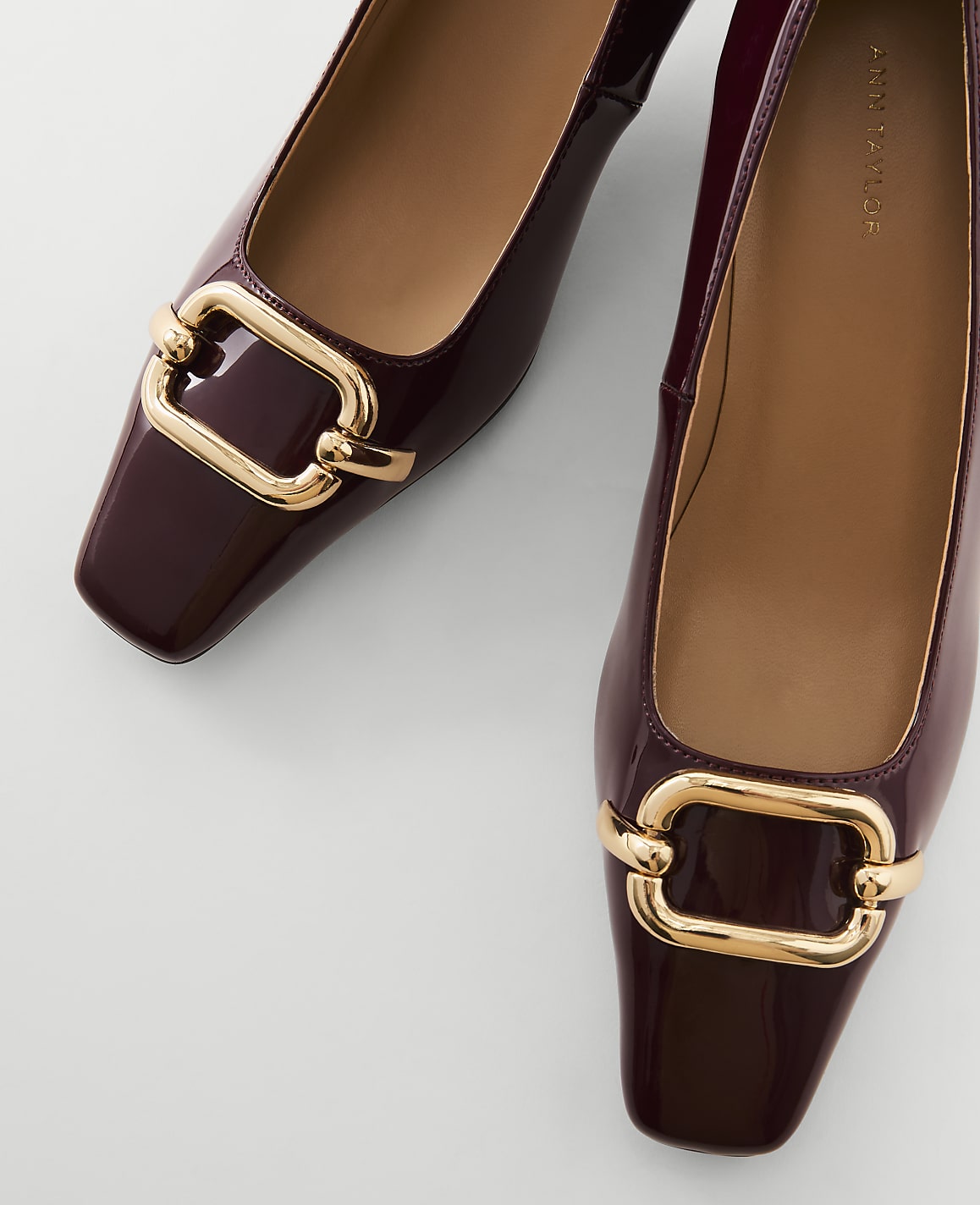 A pair of glossy burgundy square-toe flats with gold buckle details are laid out on a plain white background.