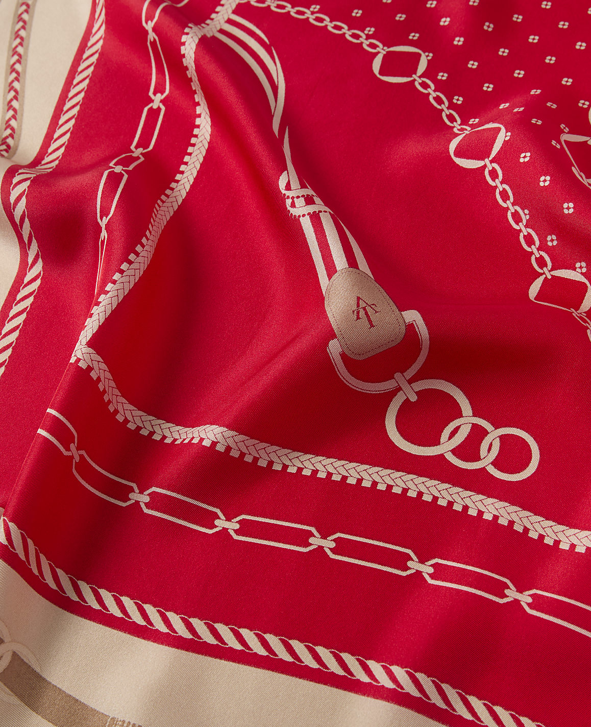 A vibrant red silk scarf with a white equestrian-themed pattern is draped in the foreground, while a blurred, neutral background provides a soft contrast.
