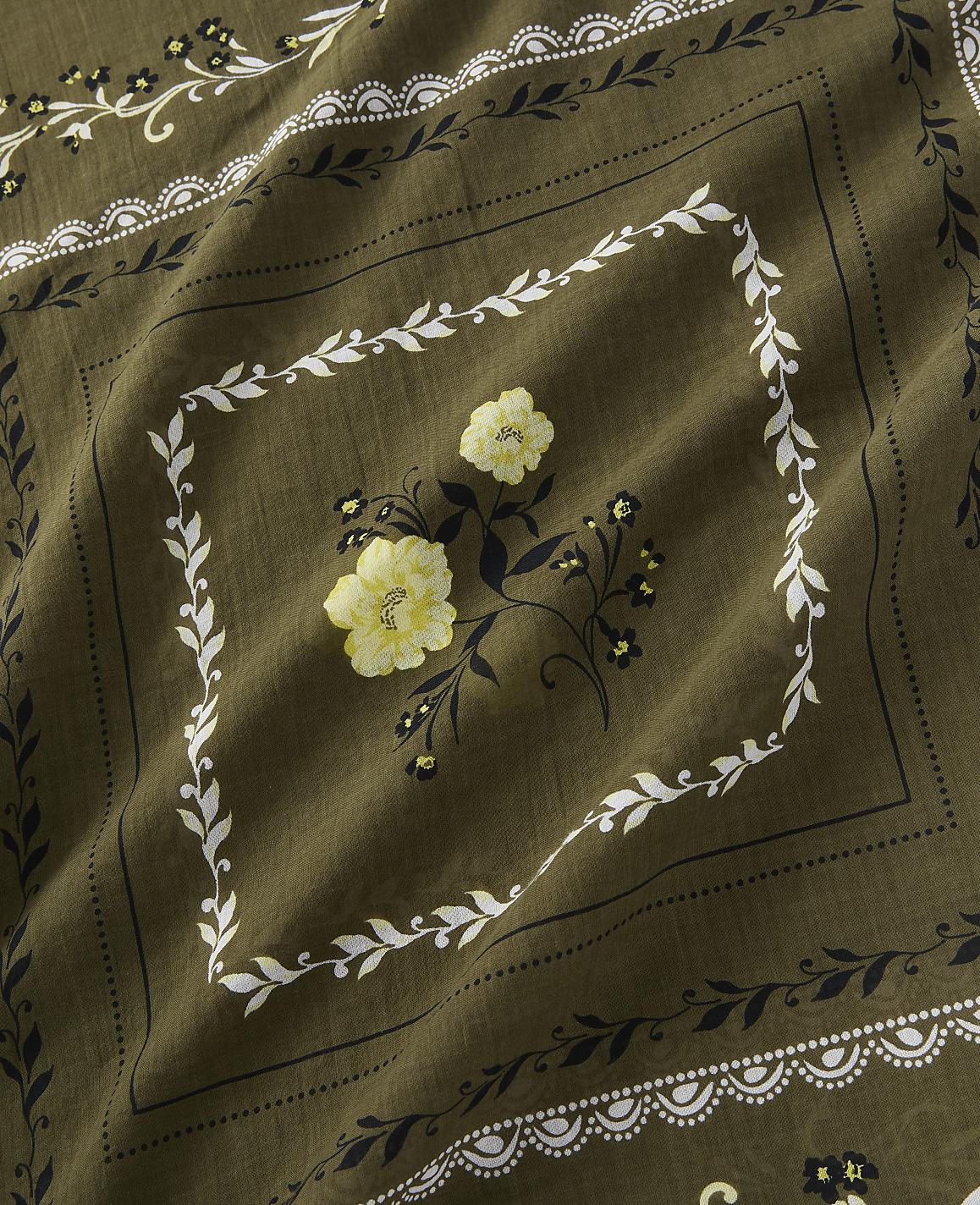 A close-up view of a khaki-colored bandana features a central diamond pattern with yellow flowers and white leafy borders, set against a textured fabric background.