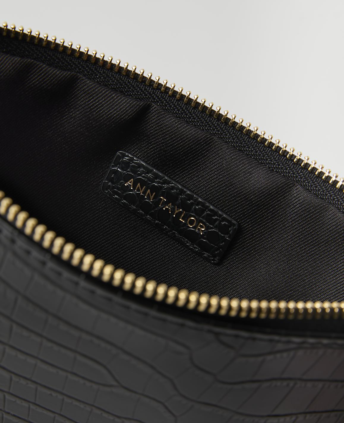 A black, crocodile-embossed clutch with a gold zipper is open to reveal a black interior with an "ANN TAYLOR" logo.