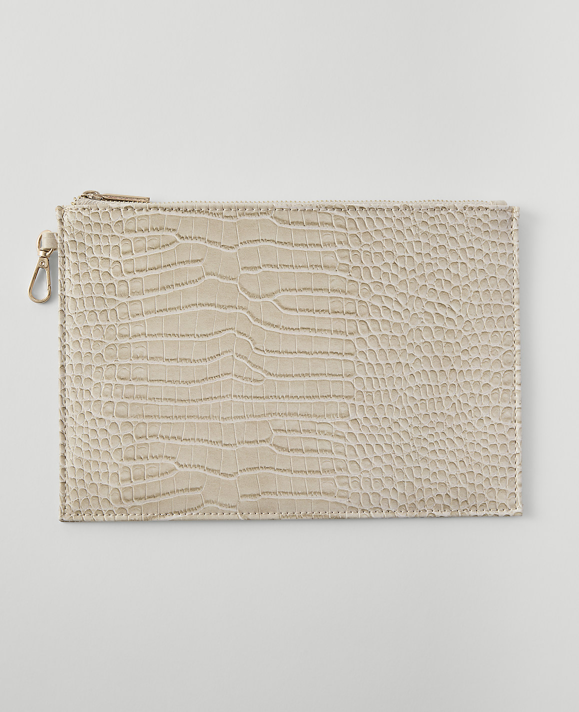 A beige, crocodile-embossed clutch with a gold zipper and clasp rests on a plain, light gray background.