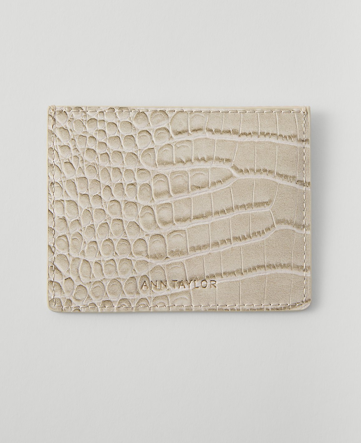 A light beige, crocodile-embossed wallet with "ANN TAYLOR" embossed on it rests on a plain white background.