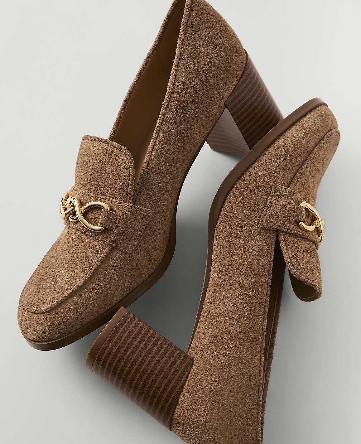 A pair of brown suede loafers with gold chain accents and chunky heels rests on a plain white background.