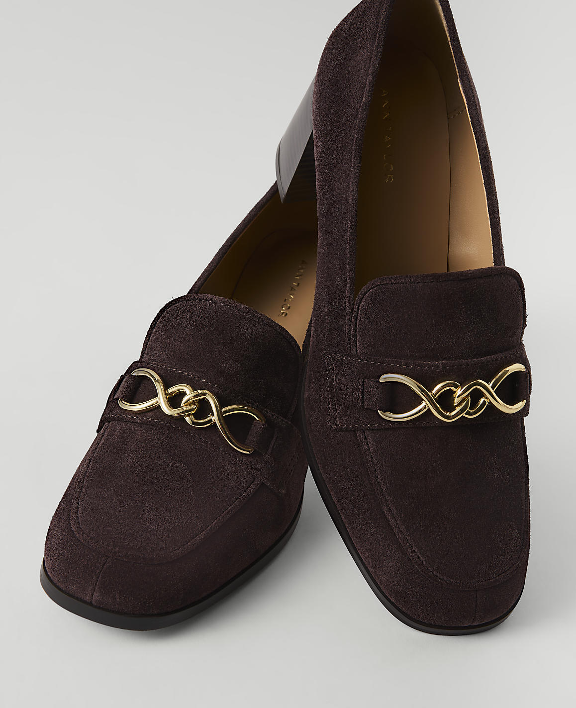 A pair of dark brown suede loafers with gold chain accents are displayed against a plain, light gray background.