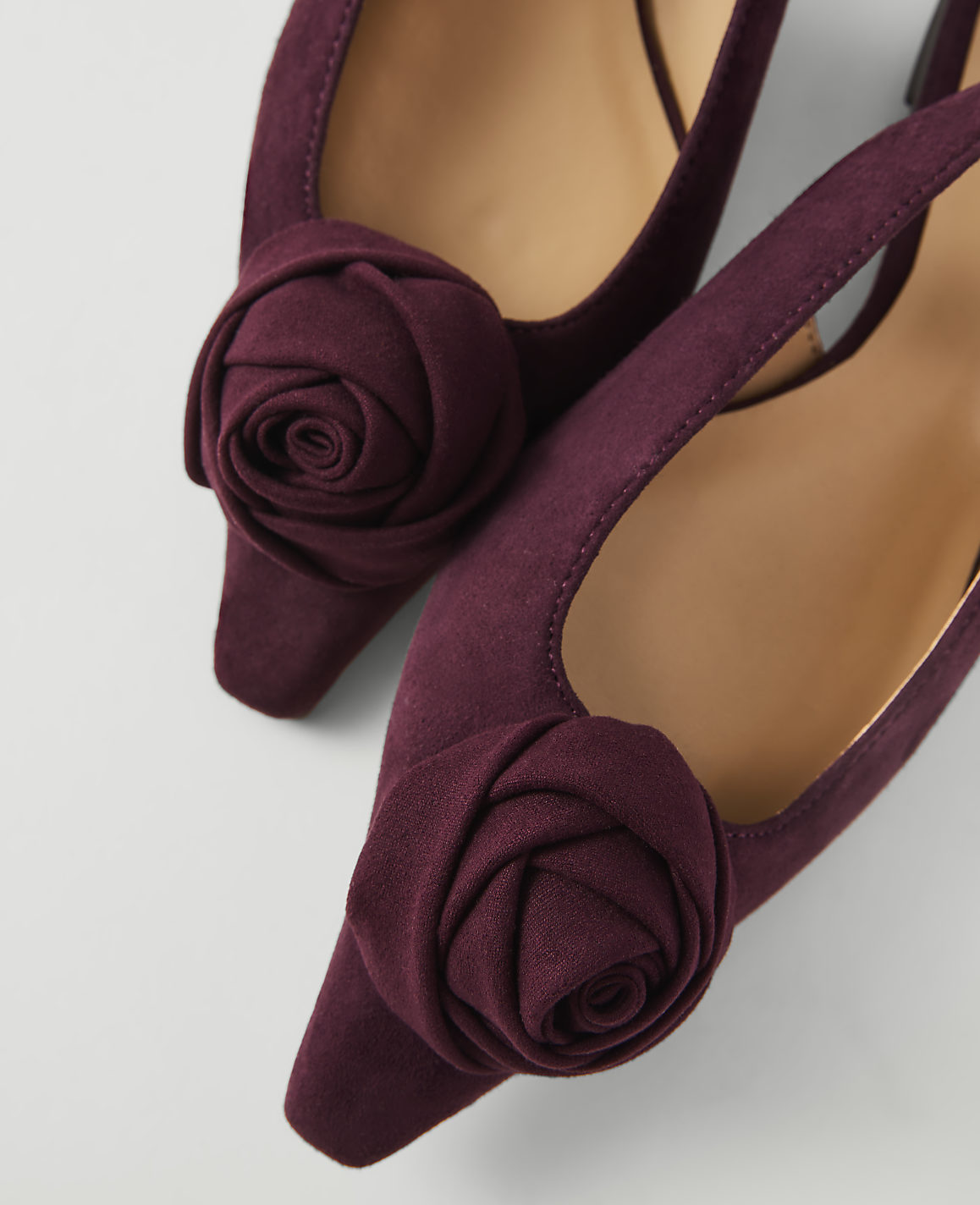 A pair of deep purple, pointed-toe flats adorned with fabric roses in the foreground are set against a plain, light gray background.