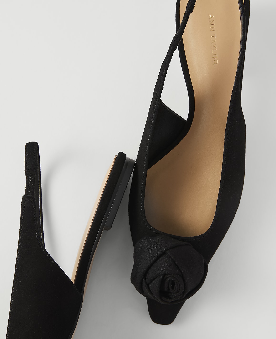 A black slingback shoe with a rose embellishment is in the foreground, with another black shoe lying beside it against a plain white background.