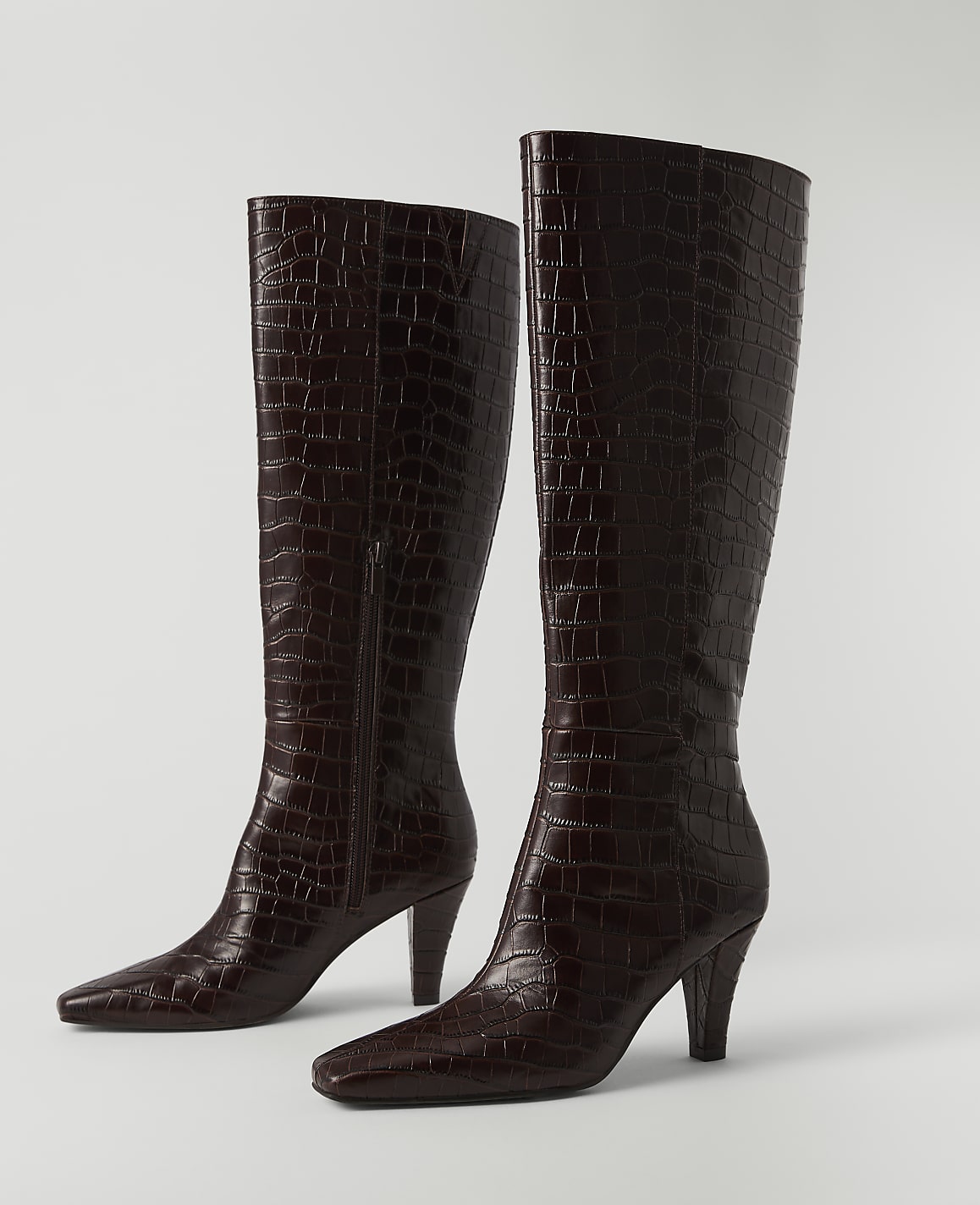 A pair of dark brown, croc-embossed leather knee-high boots with pointed toes and stiletto heels stand on a plain white surface against a white background.