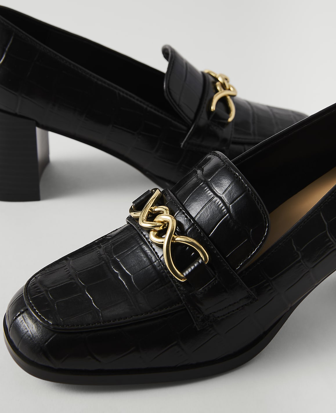 Black croc-embossed heeled loafers with gold chain accents are in the foreground, with another shoe slightly out of focus in the background.