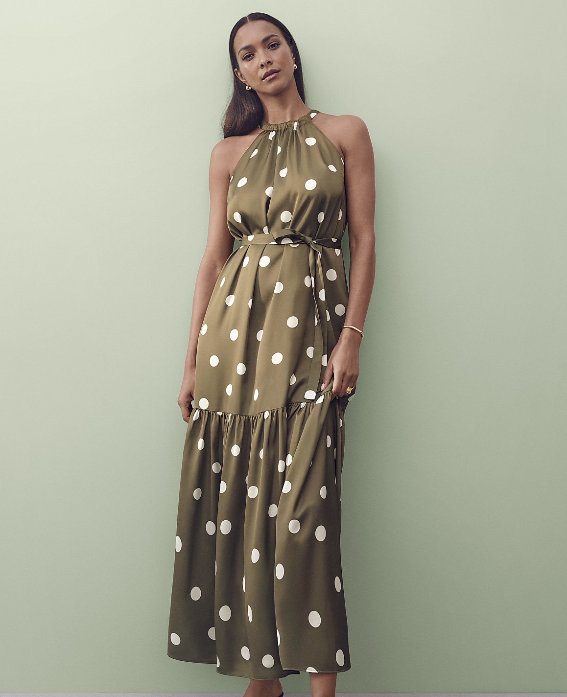 A woman in a polka-dot halter dress stands against a pale green wall.
