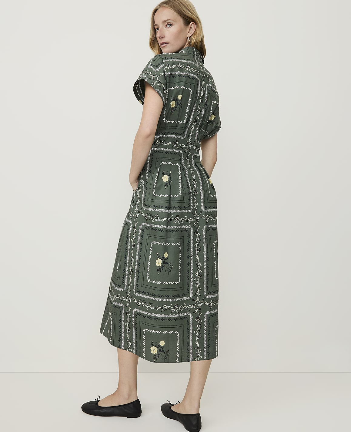 A woman in a green, patterned midi dress and black flats stands against a plain, light-colored background.