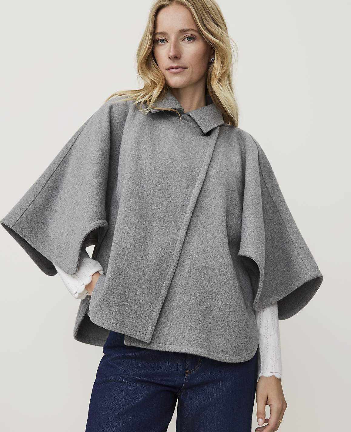 A blonde woman in a grey cape and blue jeans stands against a plain white background.