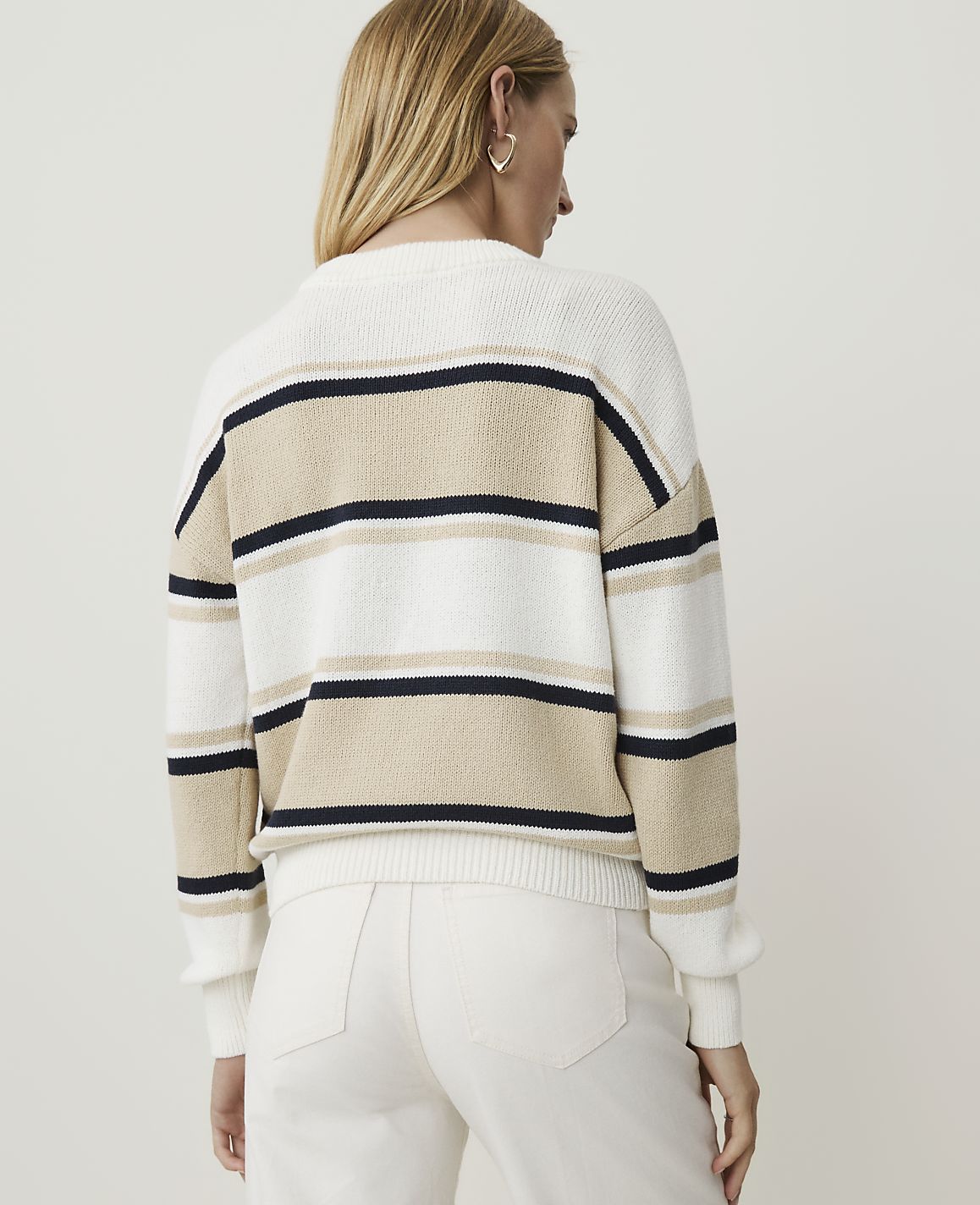 A woman in a striped sweater and white pants stands against a plain white background.
