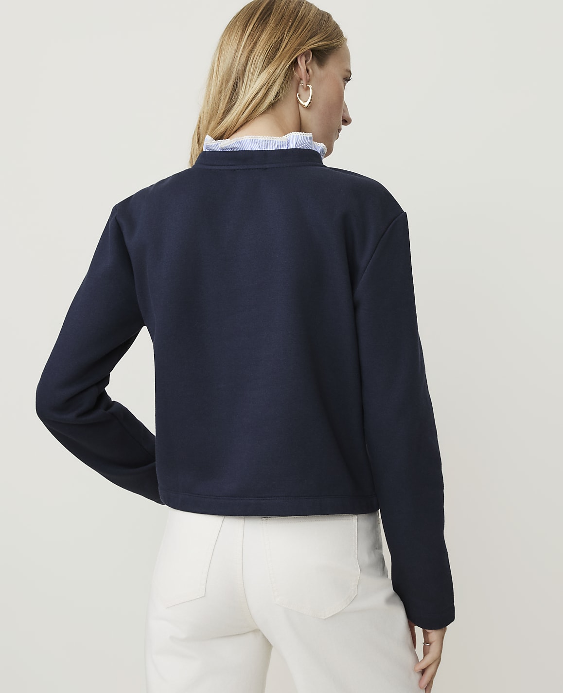 A woman in a navy sweatshirt and white pants stands against a plain white background.
