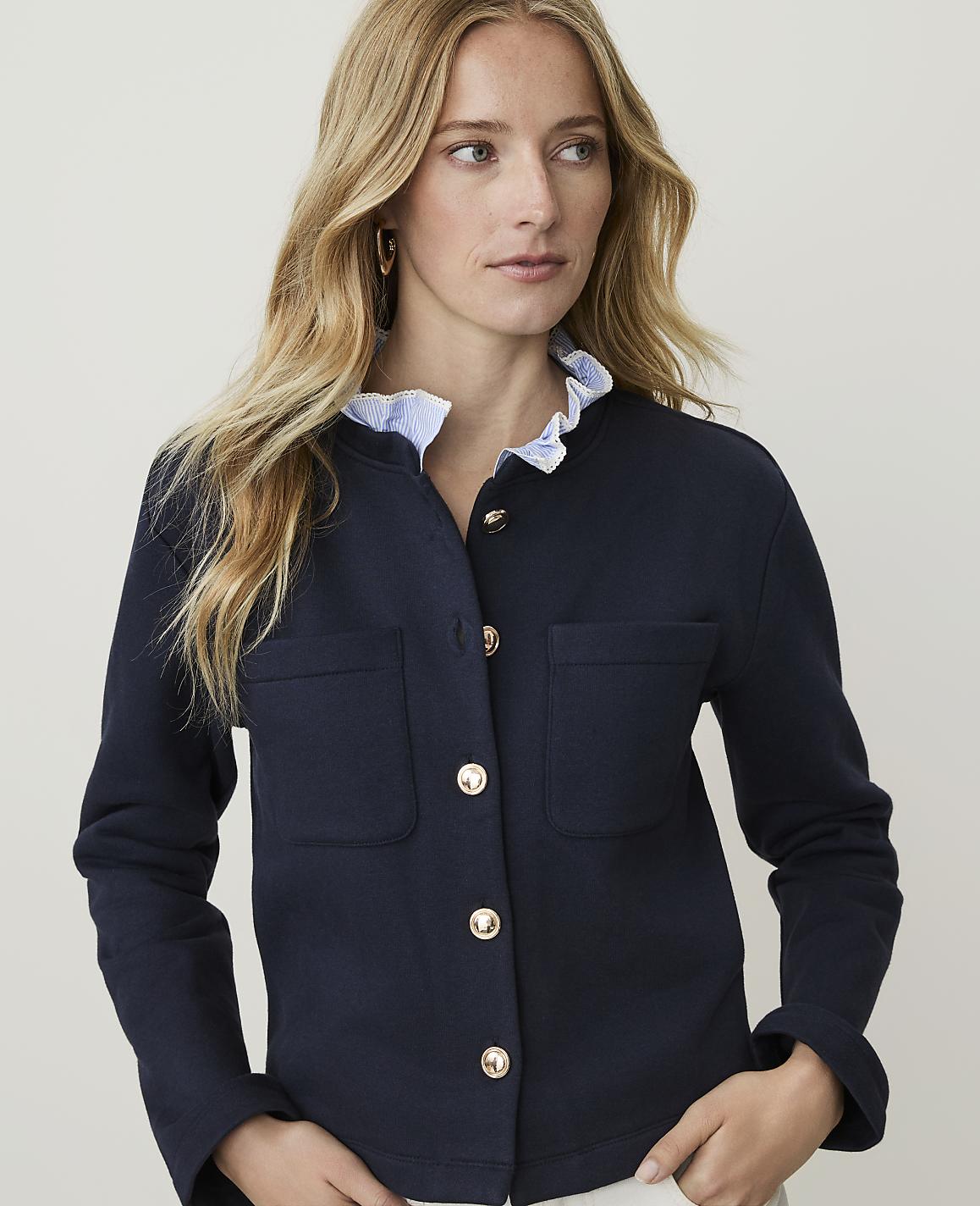 A blonde woman in a navy blue jacket with gold buttons and a ruffled collar stands against a plain white background.
