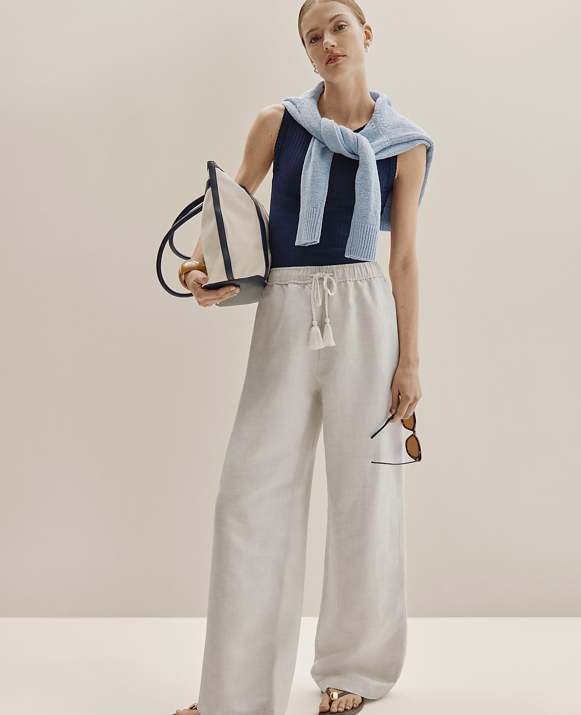 A woman in wide-leg linen pants and a striped tank top stands against a plain, light-colored background, holding a tote bag and sunglasses.