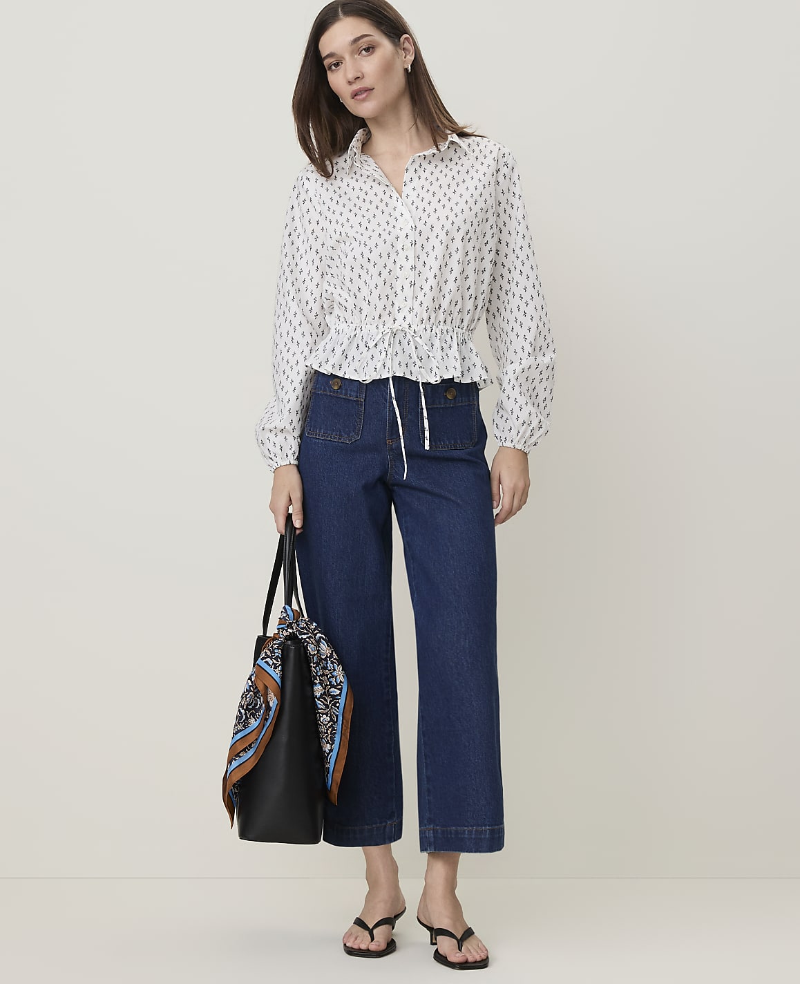 A woman in a patterned blouse and dark jeans stands with a tote bag in front of a plain white background.