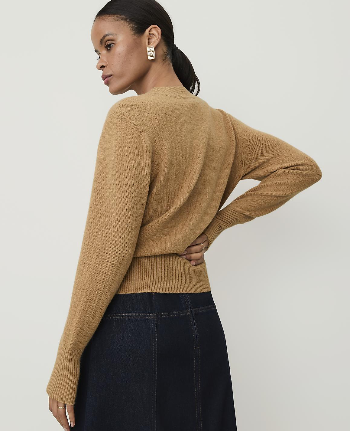 A woman in a camel-colored sweater and dark denim skirt stands against a plain white background.