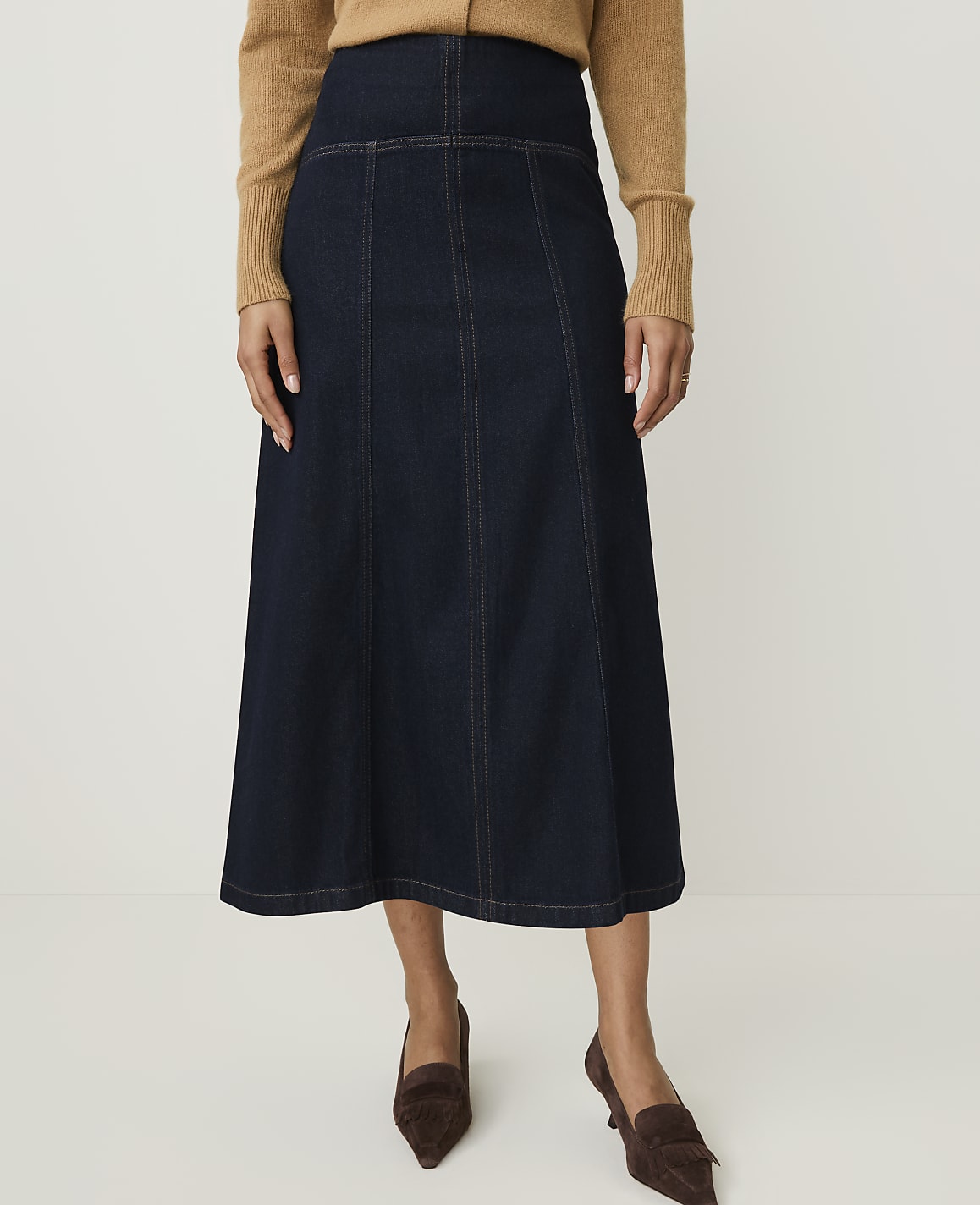 A woman in a dark denim skirt and brown sweater stands against a plain white background.