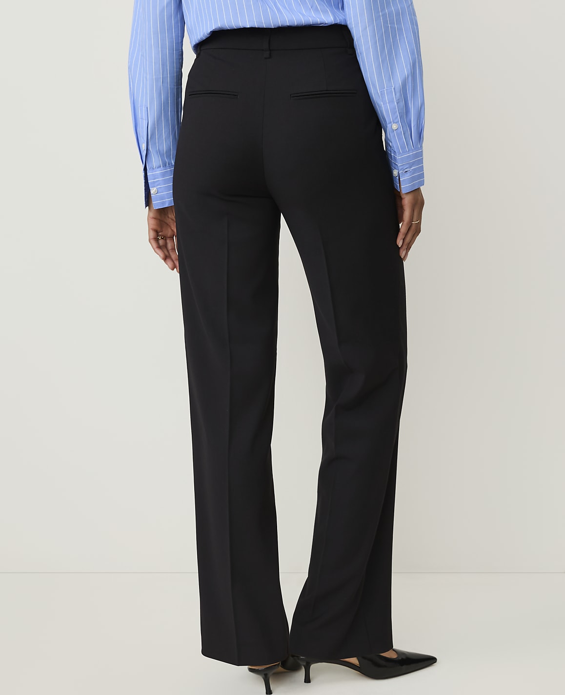 A person wearing black dress pants and a blue striped shirt stands against a plain white background.