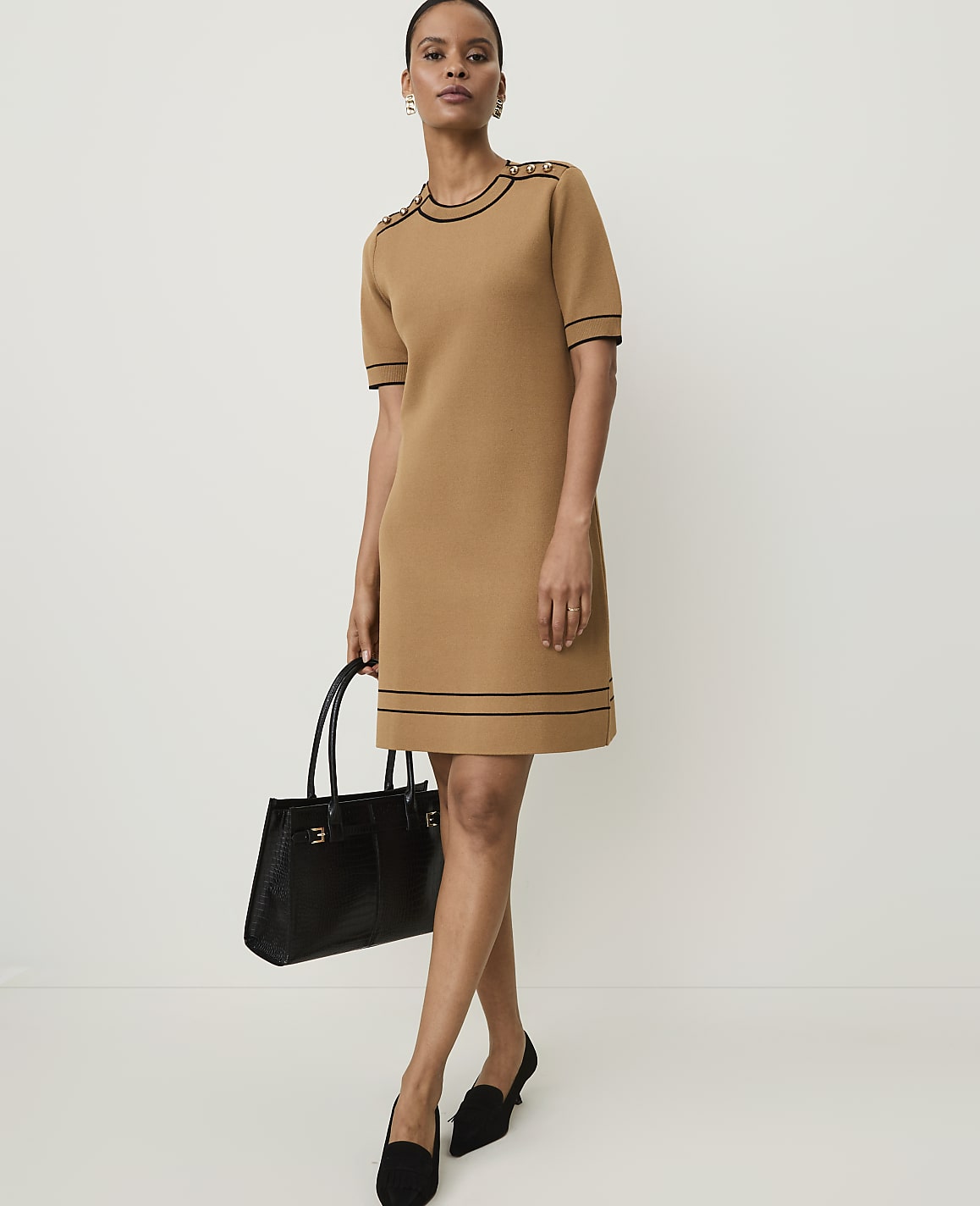 A woman in a tan knit dress with black trim stands holding a black handbag against a plain white background.