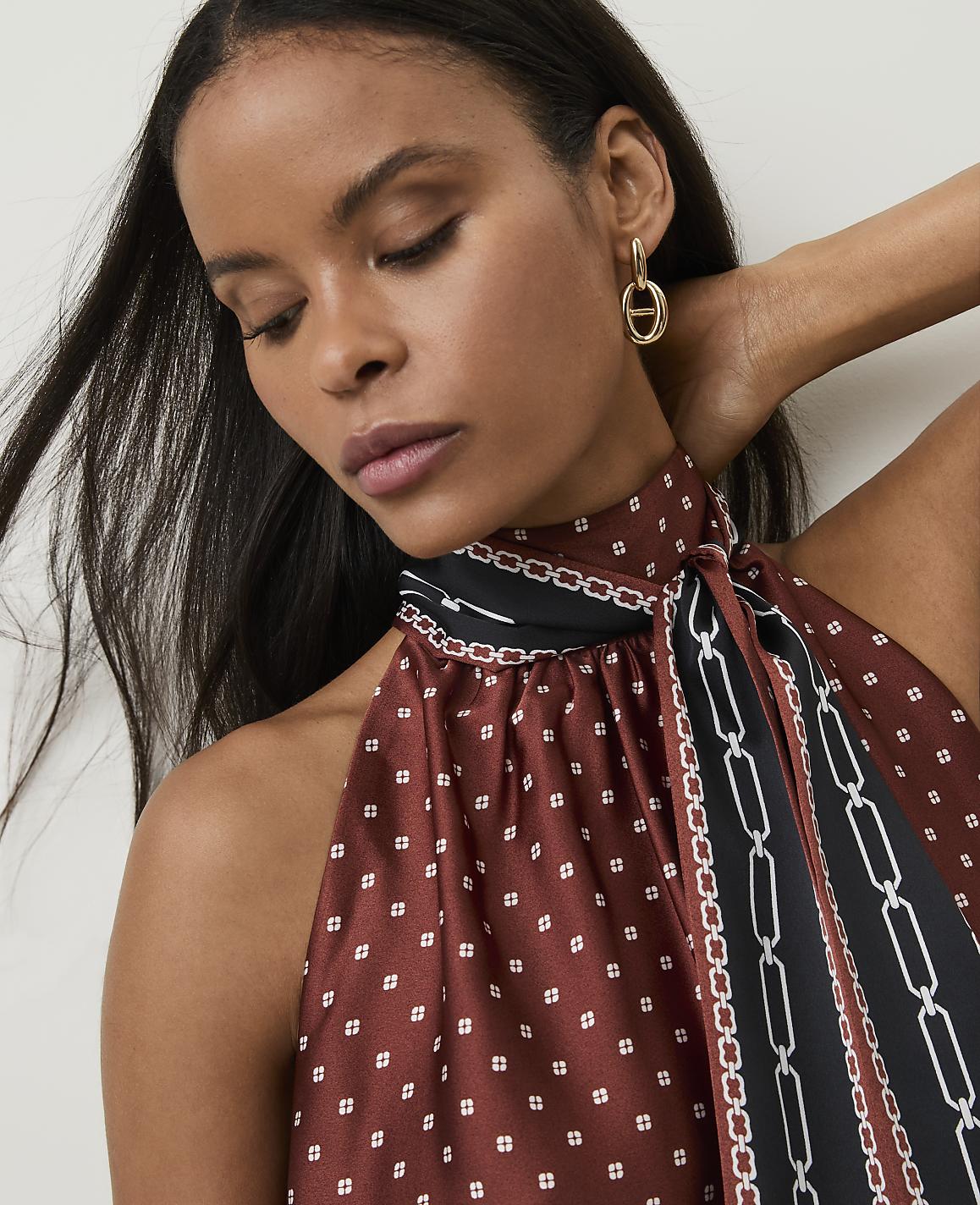 A woman with dark hair and gold earrings poses in a halter top with a patterned scarf, against a blurred background.