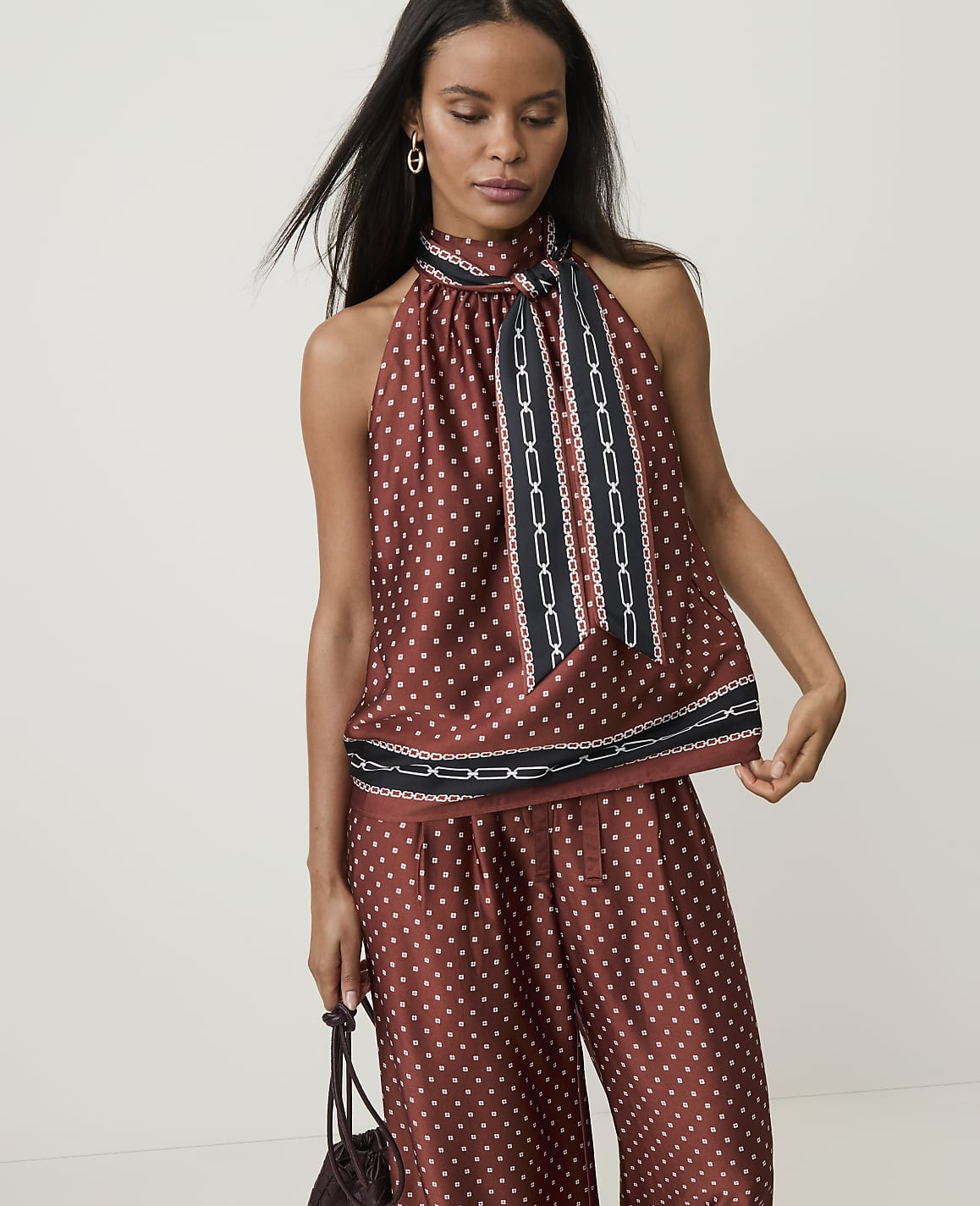 A woman in a polka-dot halter top and matching pants stands against a plain, light background, holding a dark handbag.