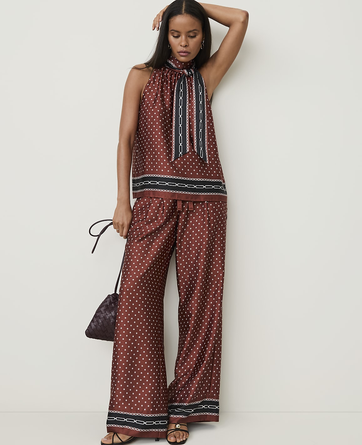 A woman in a patterned rust-colored halter top and wide-leg pants stands against a plain white background, holding a small dark purse.