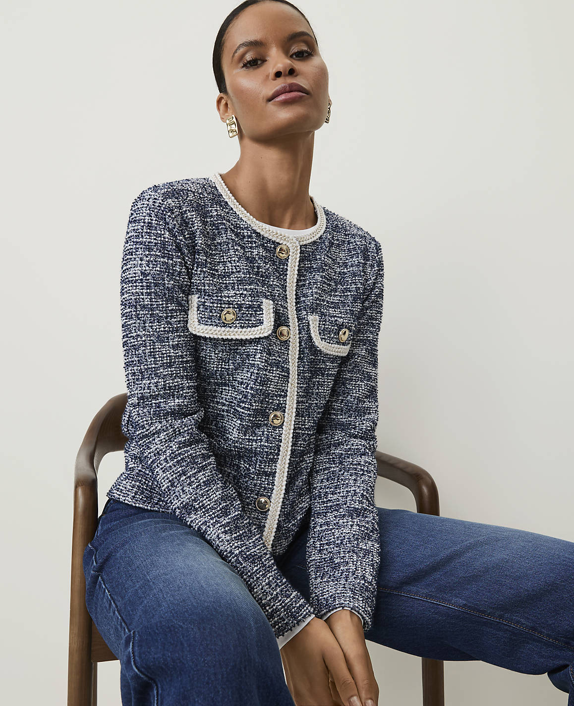 A woman in a blue tweed jacket and jeans sits on a wooden chair against a plain white background.
