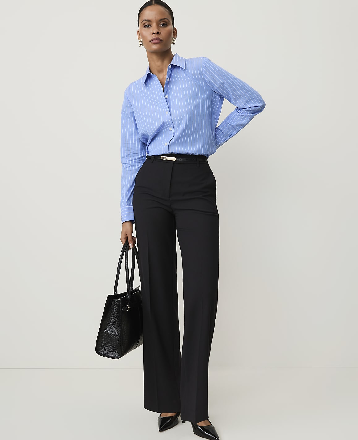 A woman in a blue pinstripe shirt and black trousers stands against a plain white background, holding a black handbag.