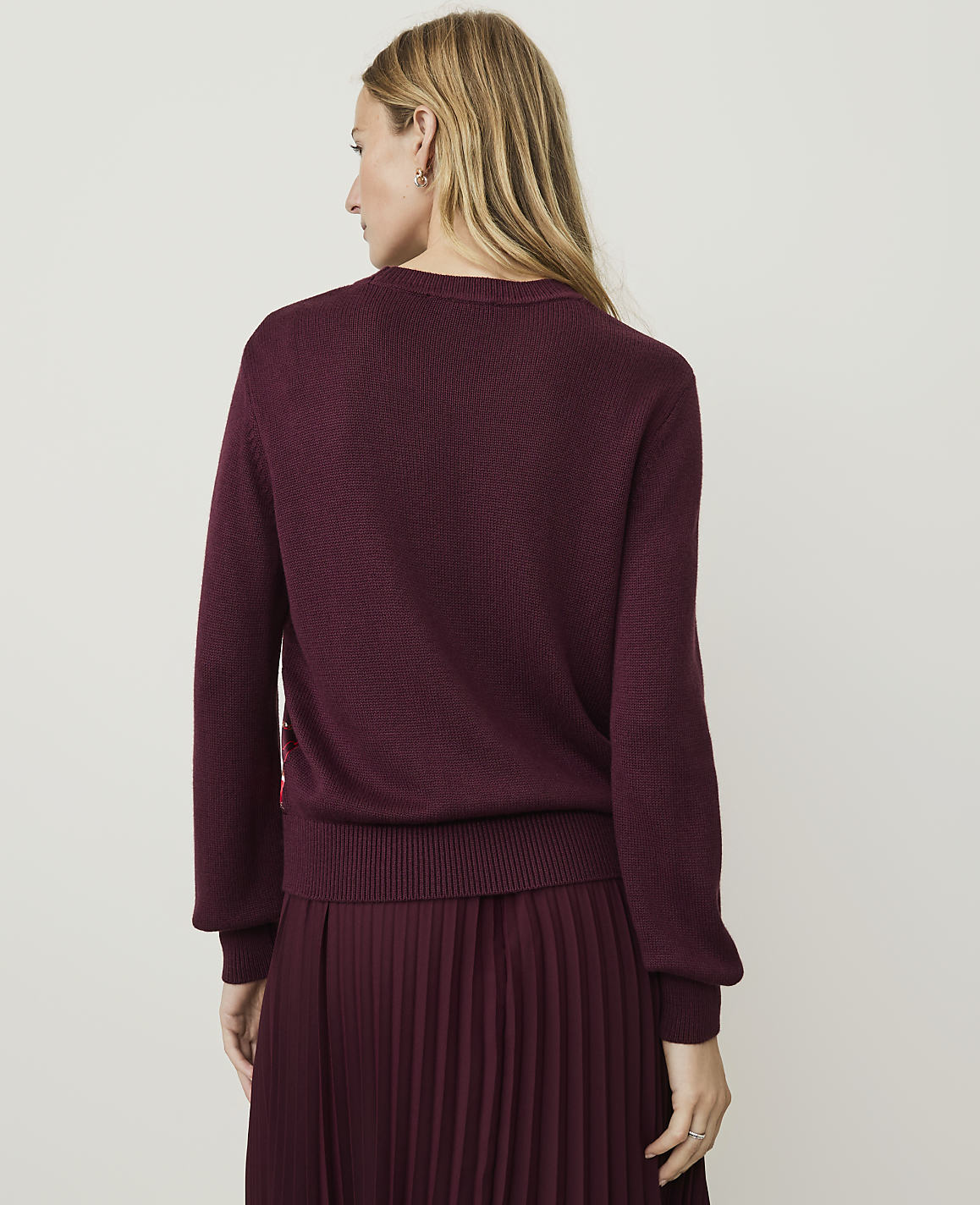 A woman in a burgundy sweater and pleated skirt stands against a plain, light-colored background.