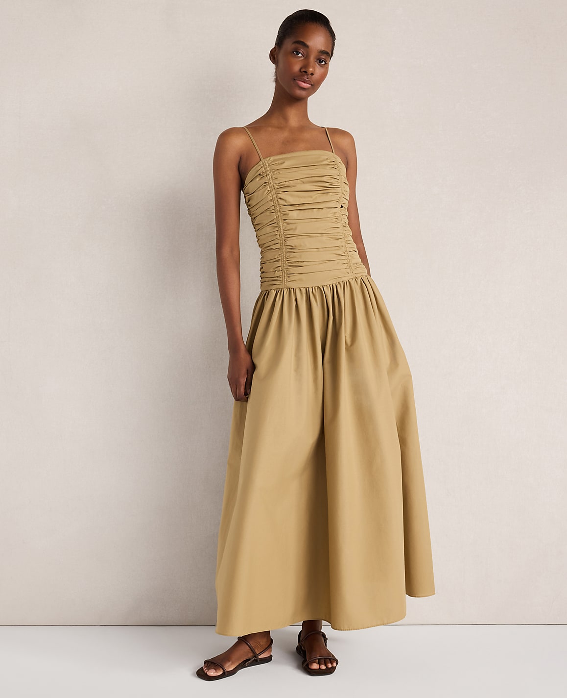 A woman in a ruched, tan maxi dress stands against a plain, light-colored wall.