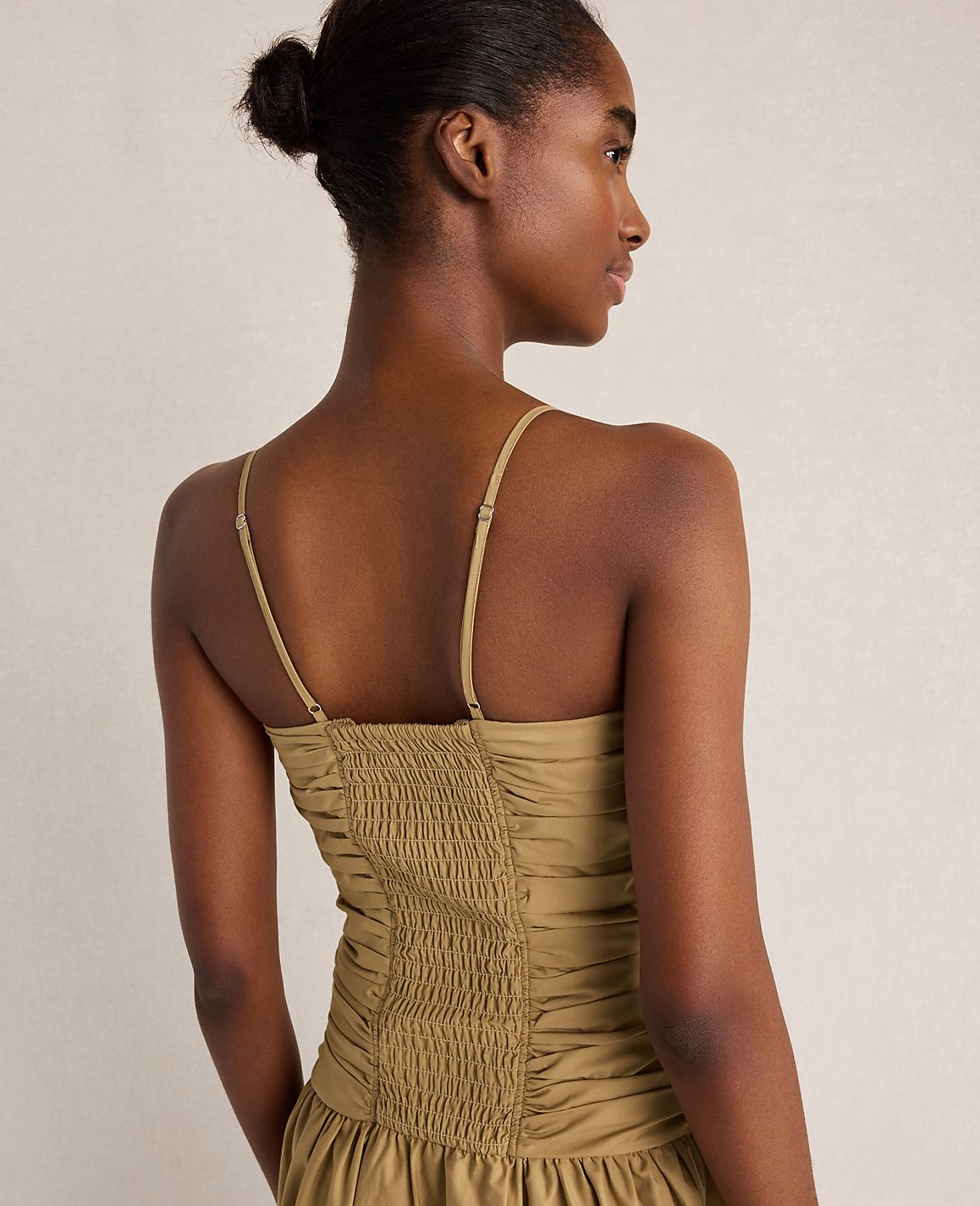 A woman in a smocked, olive-green dress is shown from the back against a plain, light-colored background.