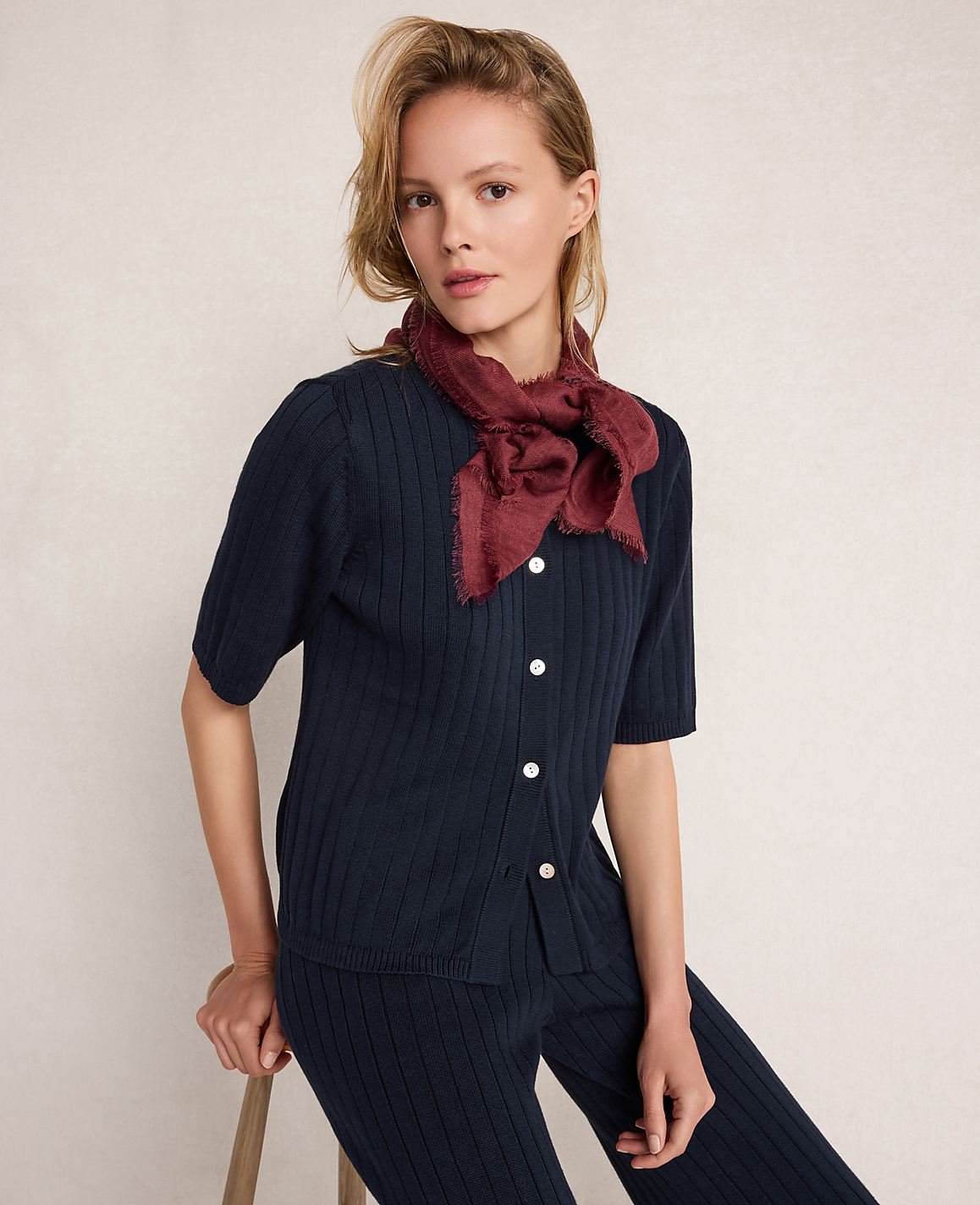 A woman in a navy ribbed outfit and a burgundy scarf sits against a plain, light background.