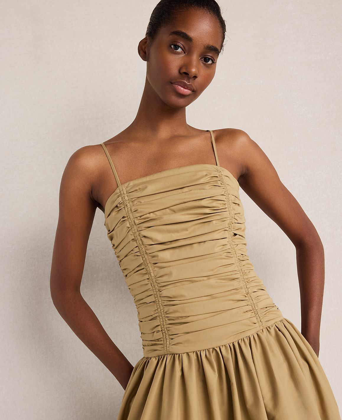 A woman in a ruched tan dress stands against a plain, light-colored background.