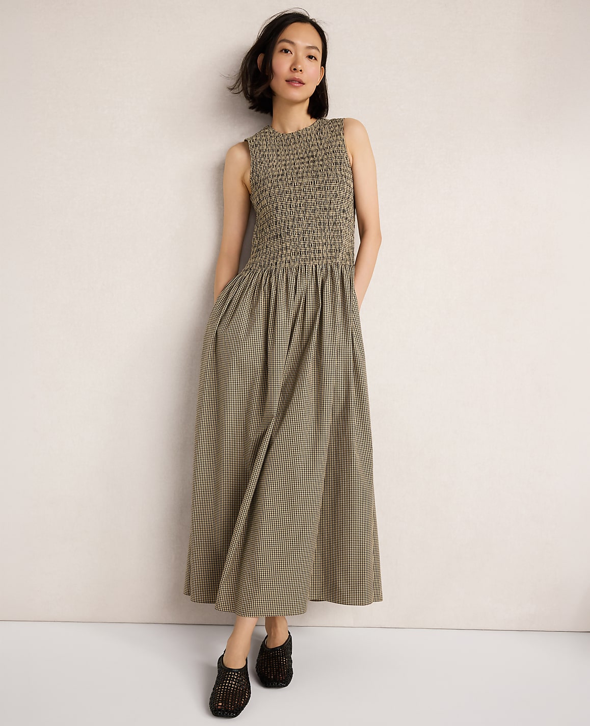 A woman in a smocked olive green dress stands against a plain, light-colored wall.